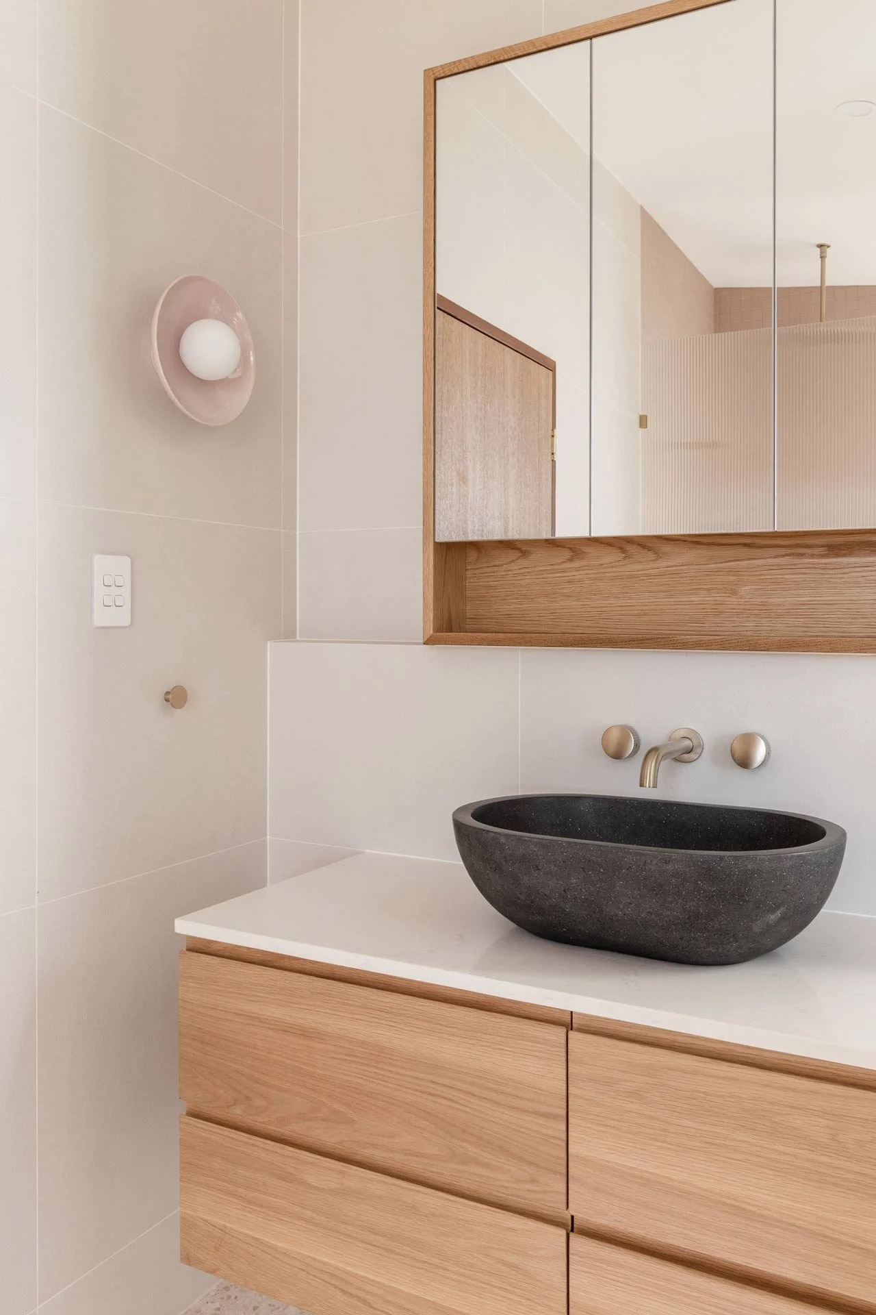 Modern bathroom with a black stone sink on a white countertop, light wooden vanity, mirrored cabinet, and beige wall tiles.