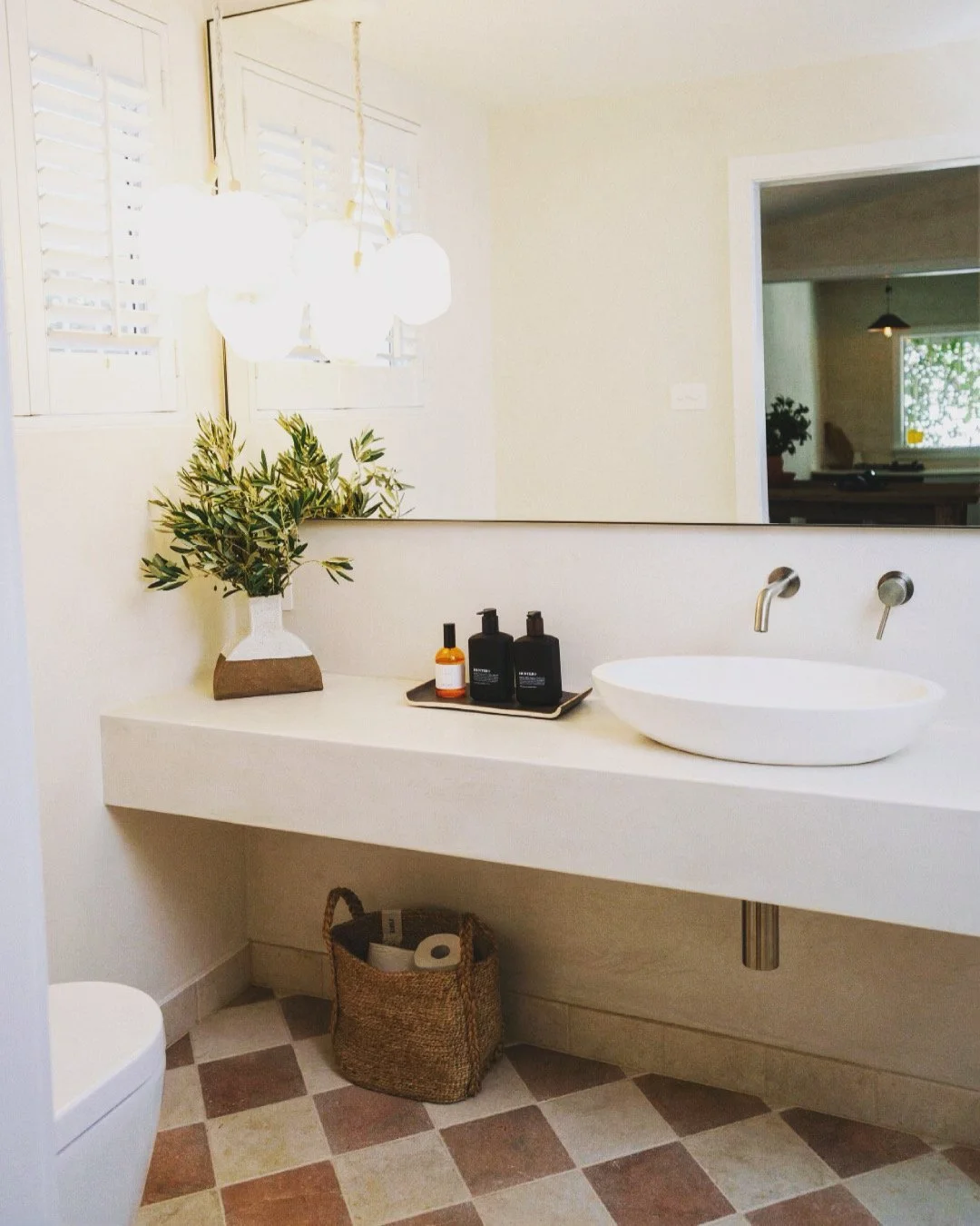 Modern bathroom with a floating white vanity, oval vessel sink, and wall-mounted faucet. Decor includes a potted plant, black soap dispensers, and a woven basket with toilet paper. There are windows with white shutters and a large mirror above the vanity.