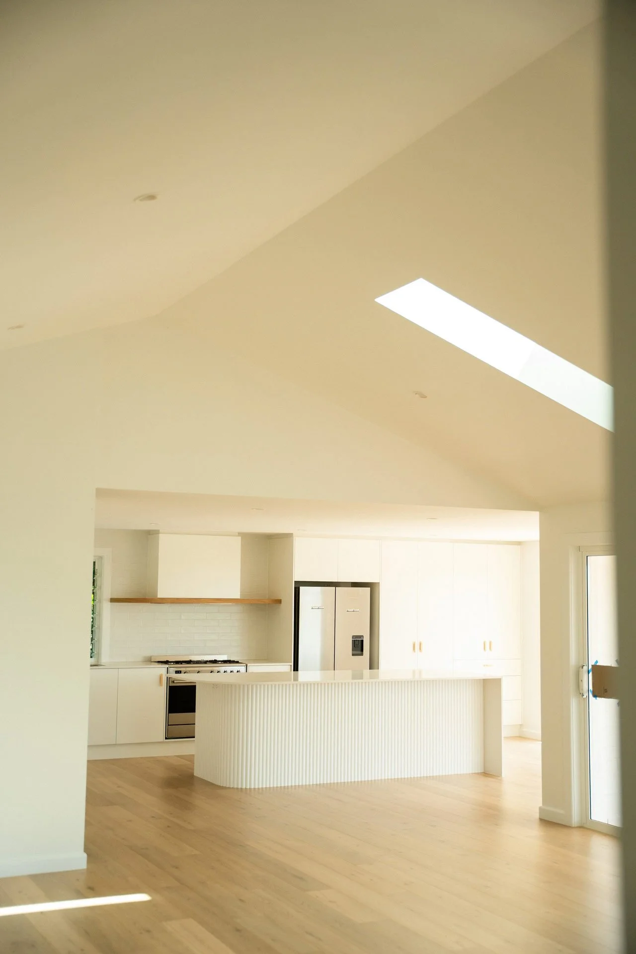 Open-concept kitchen with white cabinets, a stainless steel refrigerator, and a white kitchen island with vertical ridges, illuminated by natural light from large windows.