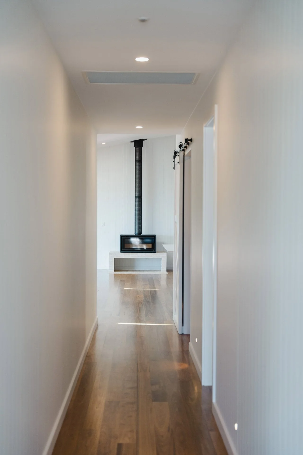 A narrow hallway with white walls, wooden flooring, and ceiling lights leading to a room with a modern fireplace and a black chimney pipe.