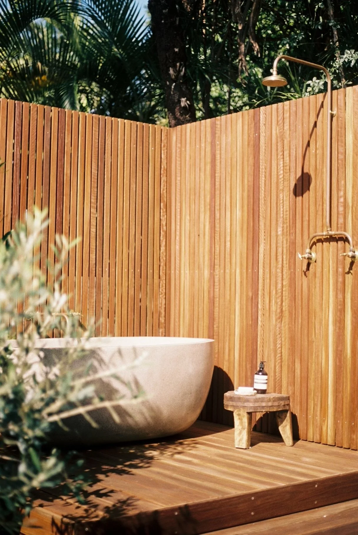 Outdoor shower area with a wooden privacy fence, a freestanding bathtub, and a small wooden stool with soap, lotion, and a towel. Lush greenery visible behind the fence.