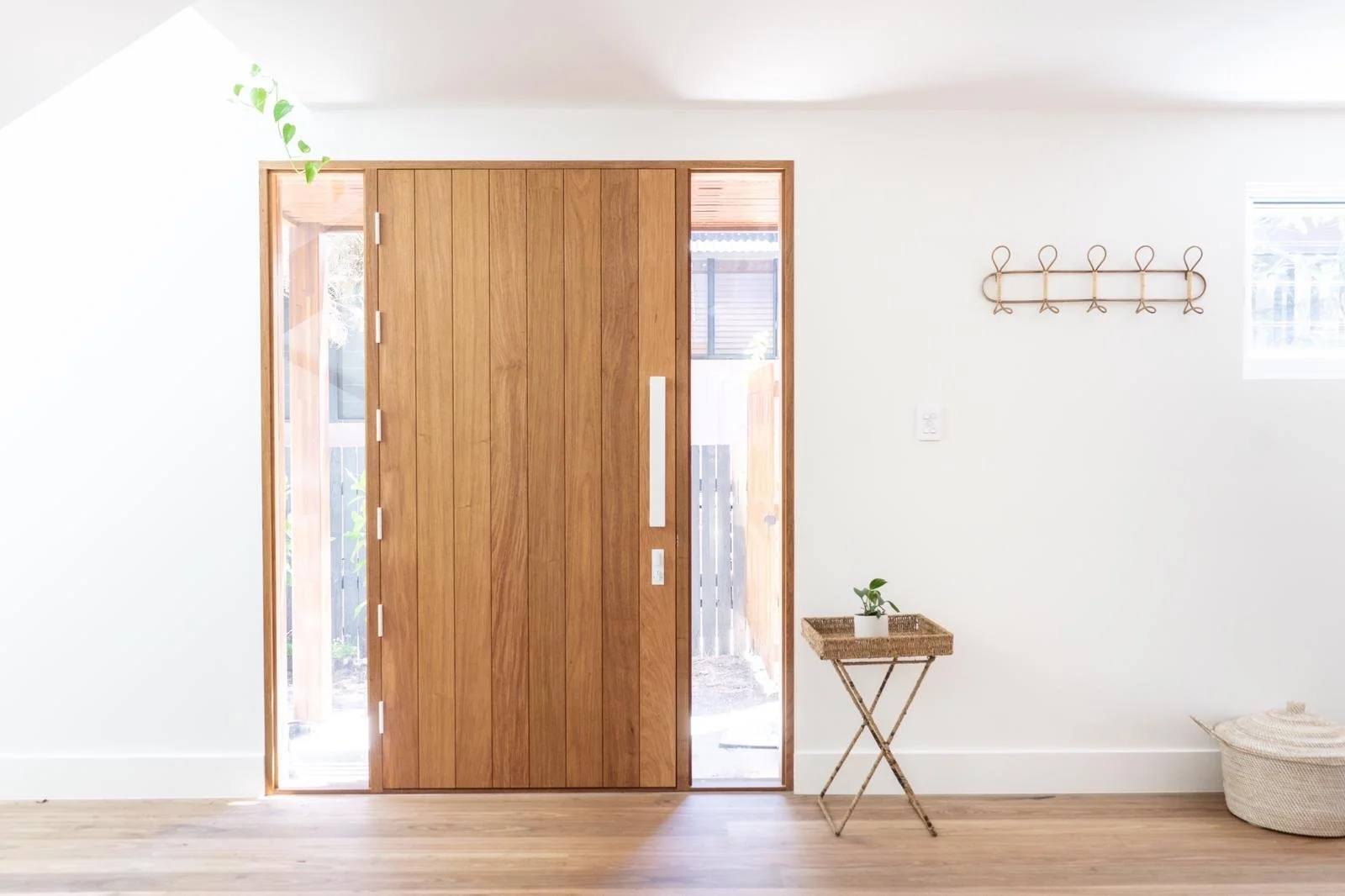 Interior view of an entryway with a wooden door and side windows, white walls, a woven side table with a small plant, a wall-mounted hook rack, a woven basket, and hardwood flooring.