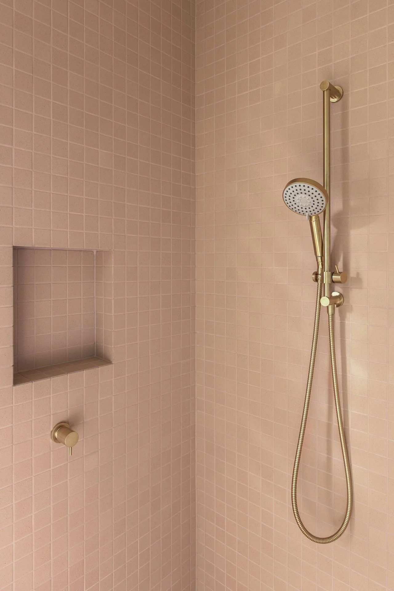 A walk-in shower with pink square tiles, a niche shelf, and a gold-finish showerhead with a handheld attachment.
