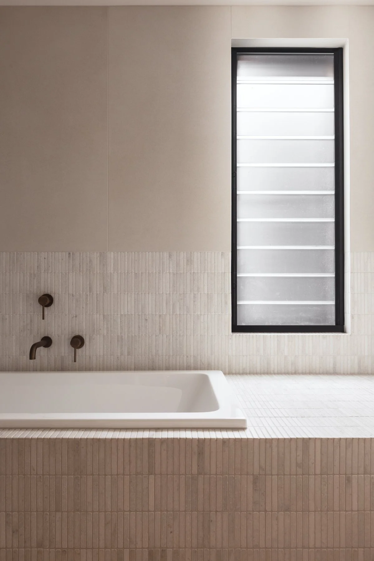 Bathroom with a rectangular bathtub, beige tile walls, and a tall, narrow window with frosted glass and horizontal lines.