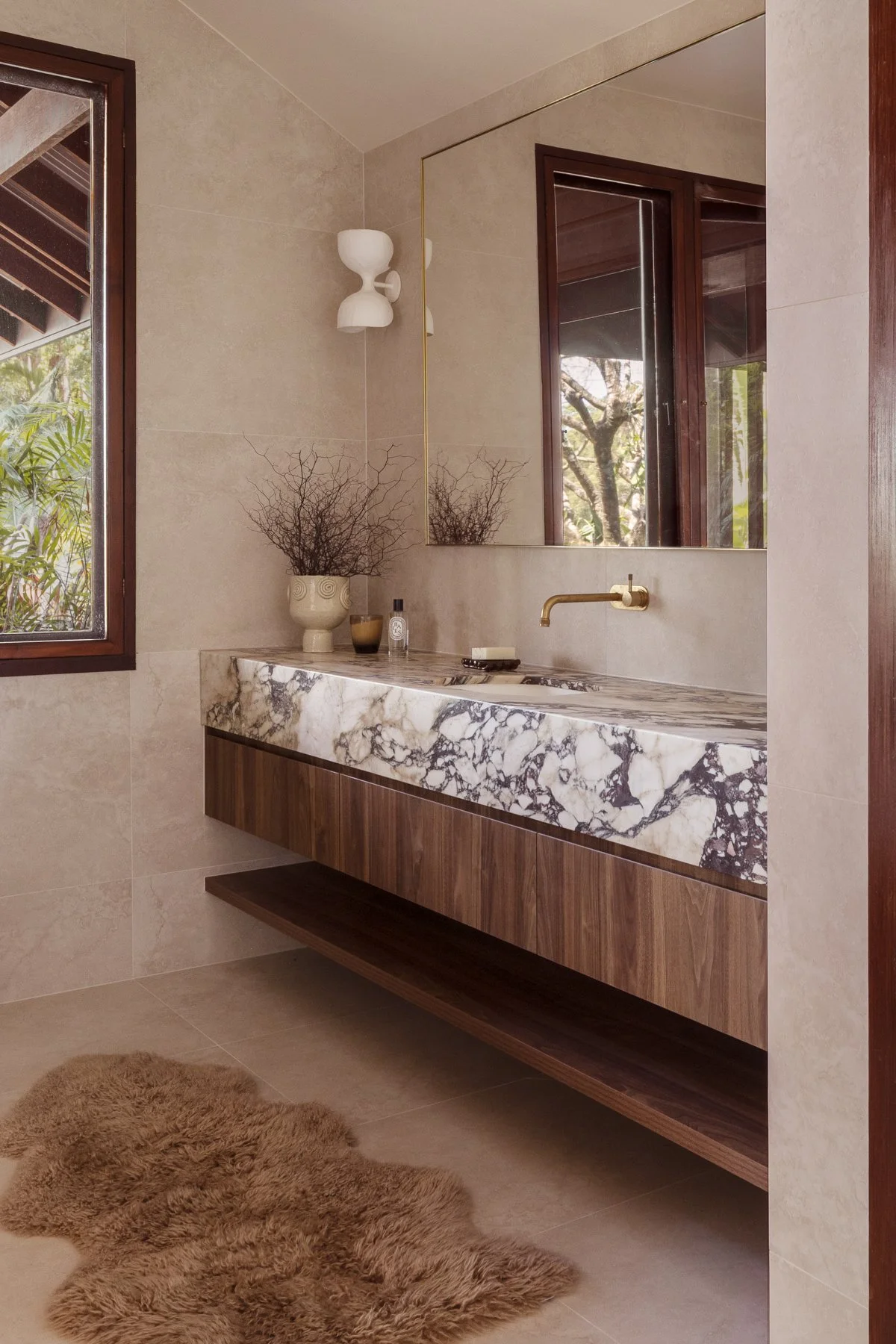 Modern bathroom with beige tiled walls and floor, featuring a floating marble vanity with a dark wood base, a gold wall-mounted faucet, a large rectangular mirror, a window with views of trees, a white decorative vase with branches, and a fluffy beige rug on the floor.