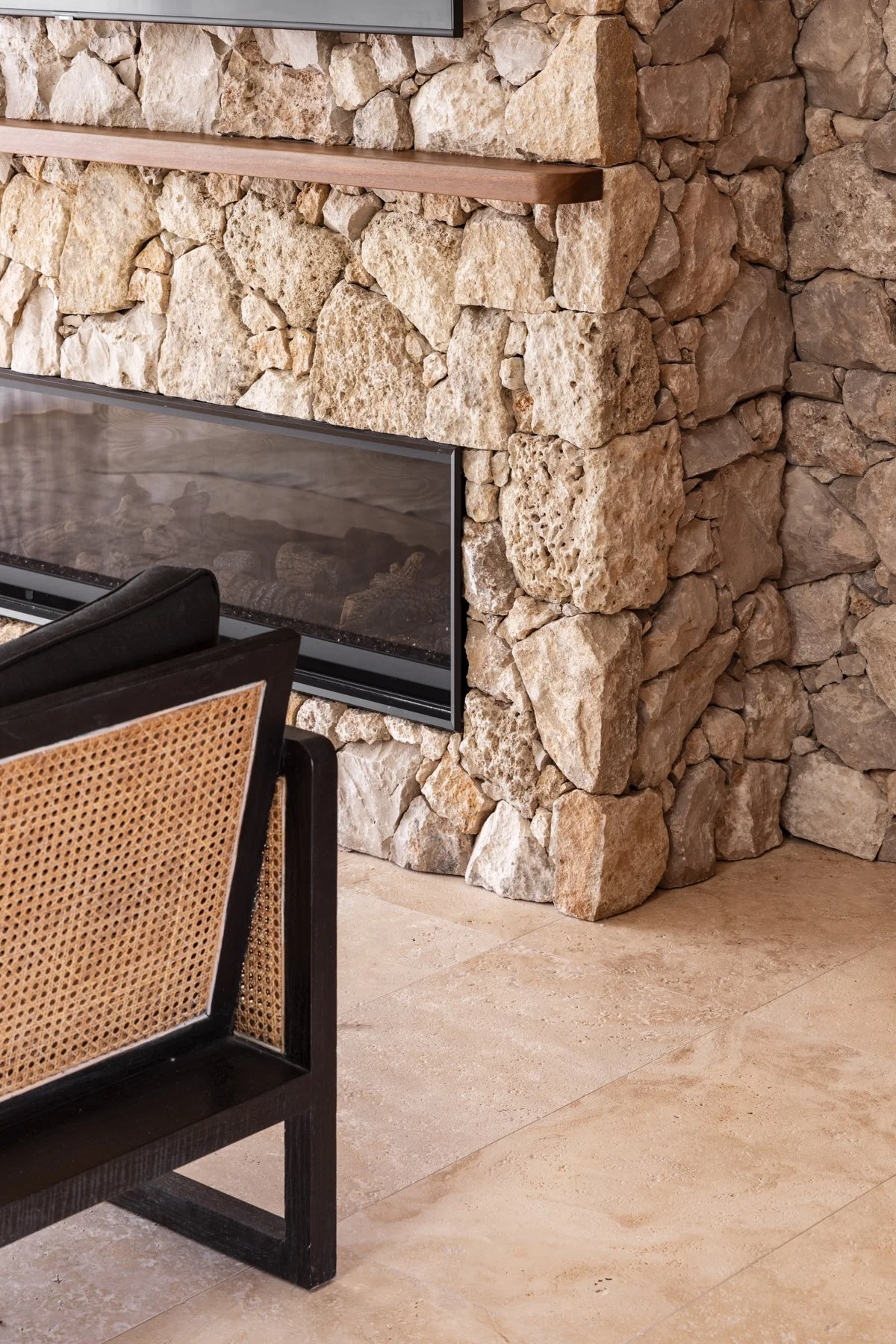 Close-up view of a fireplace with a stone facade, a glass firebox, and a wooden mantel in a living room with stone tiled flooring.