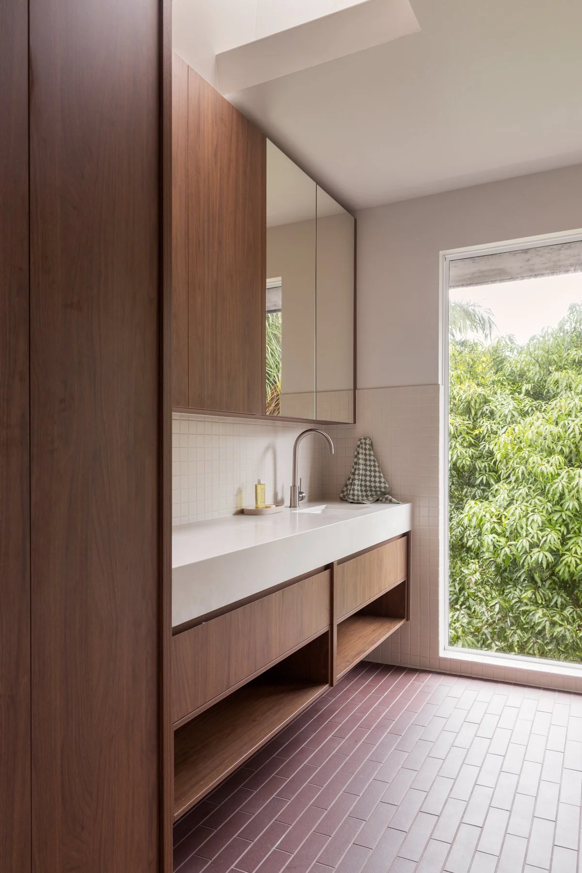 A bathroom with wooden cabinetry, a white countertop with a built-in sink, a chrome faucet, and a large window with greenery outside.