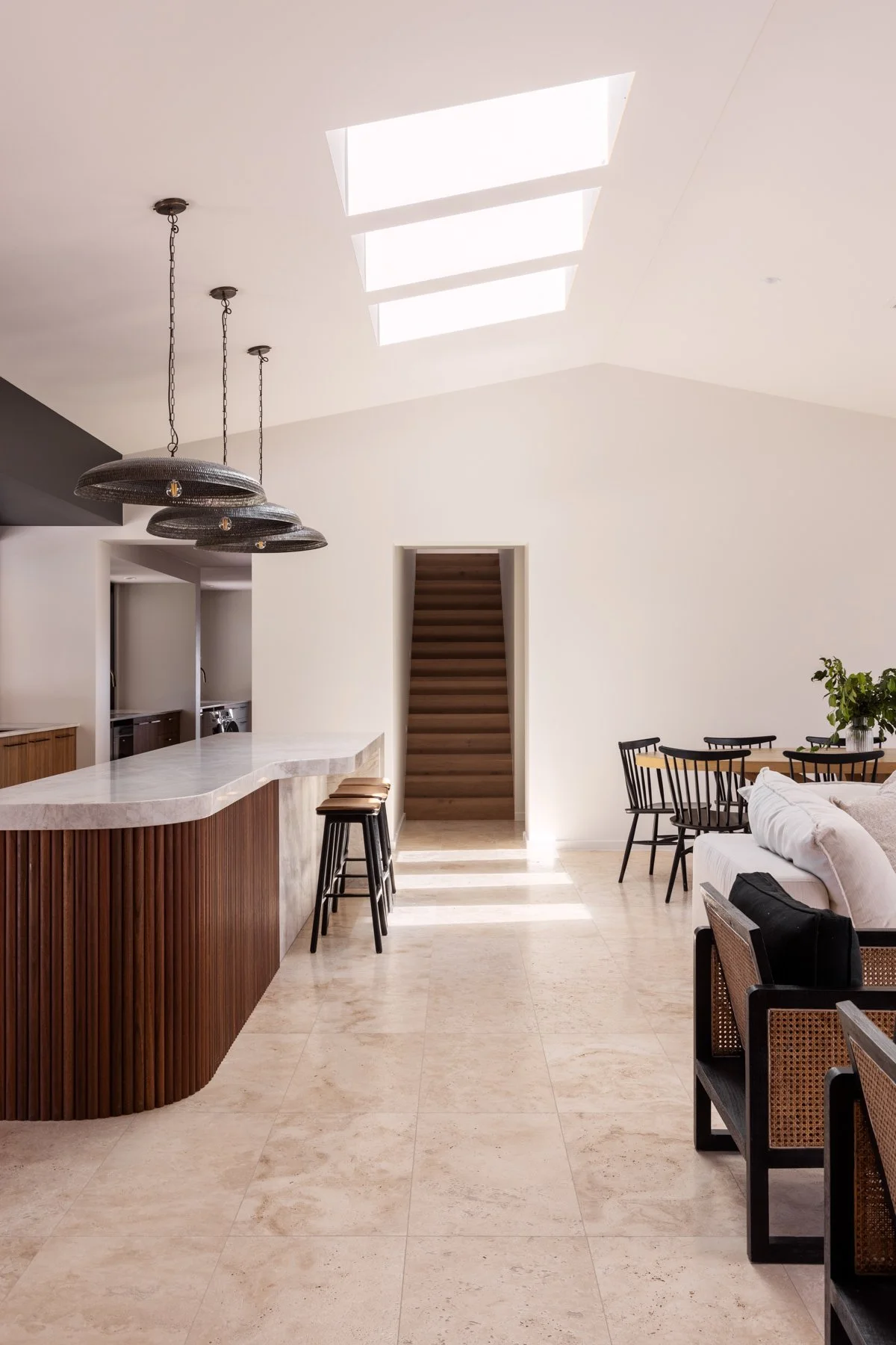 Modern interior dining and kitchen area with a high ceiling, skylights, a marble-topped kitchen island with a wooden base, black bar stools, black dining chairs with light-colored seats, a split-level staircase in the background, and minimal decoration including a potted plant on the dining table.