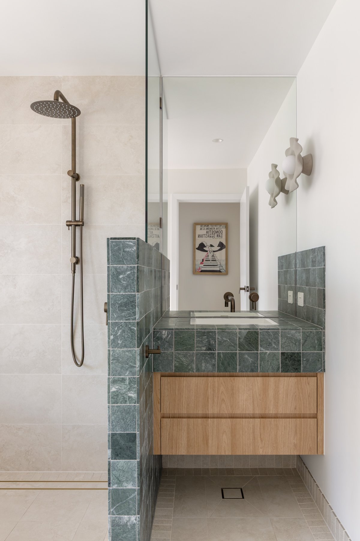 Modern bathroom with walk-in shower on the left, featuring a large showerhead and a handheld shower. To the right, a green tiled vanity with a built-in sink and a wooden cabinet beneath. Above, white walls with decorative wall sconces. In the background, a framed artwork hanging on the wall opposite the vanity, visible through a doorway.