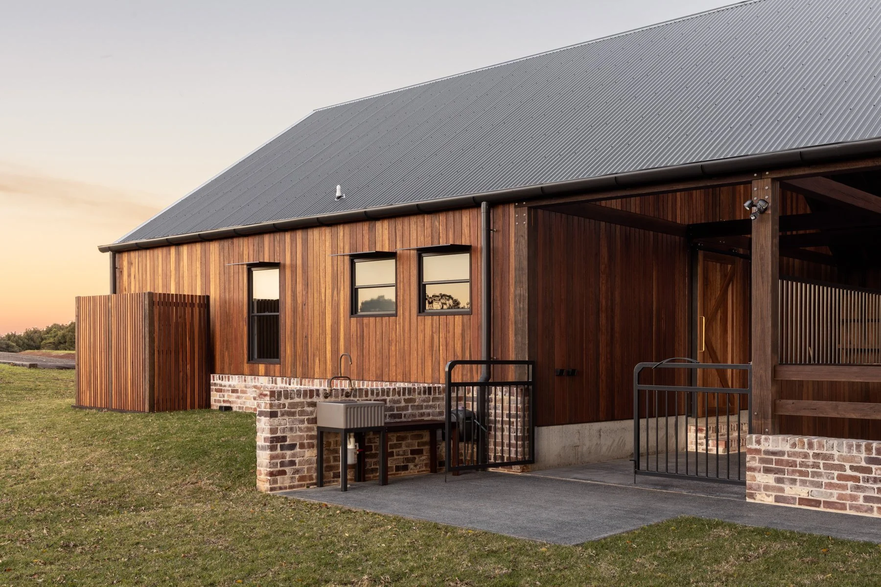 A modern building exterior with wooden siding, black-framed windows, and a gray metal roof. There are outdoor utilities including a utility sink and a small fenced area, with a grassy yard and a sunset sky in the background.