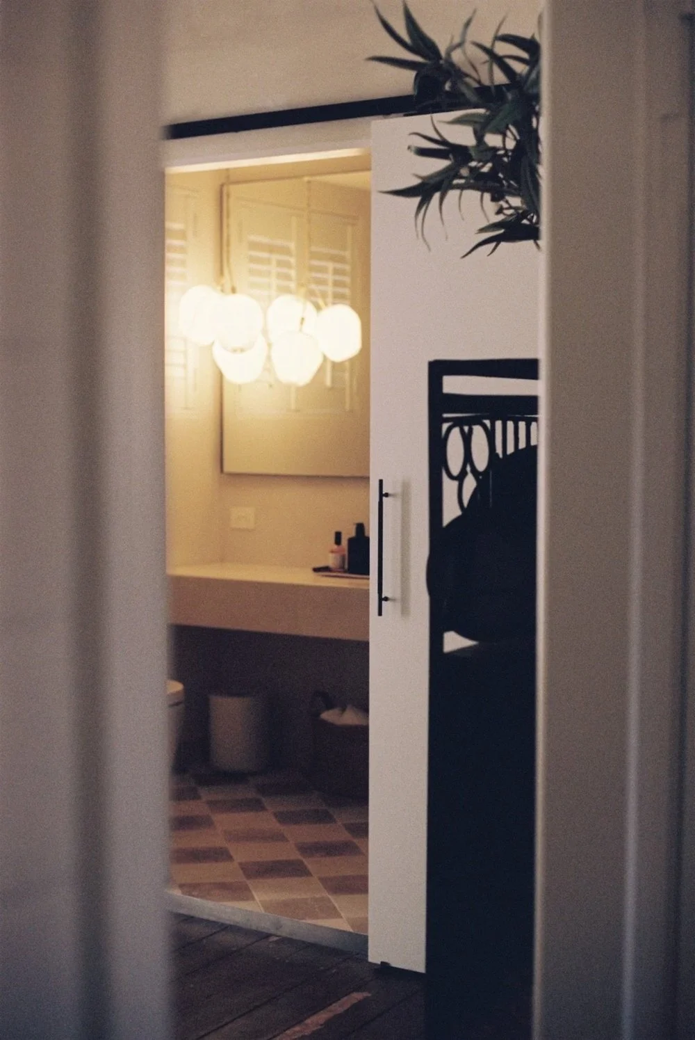 Interior view of a bathroom seen through partially opened door, with a glowing ceiling light fixture, mirror, and vanity.