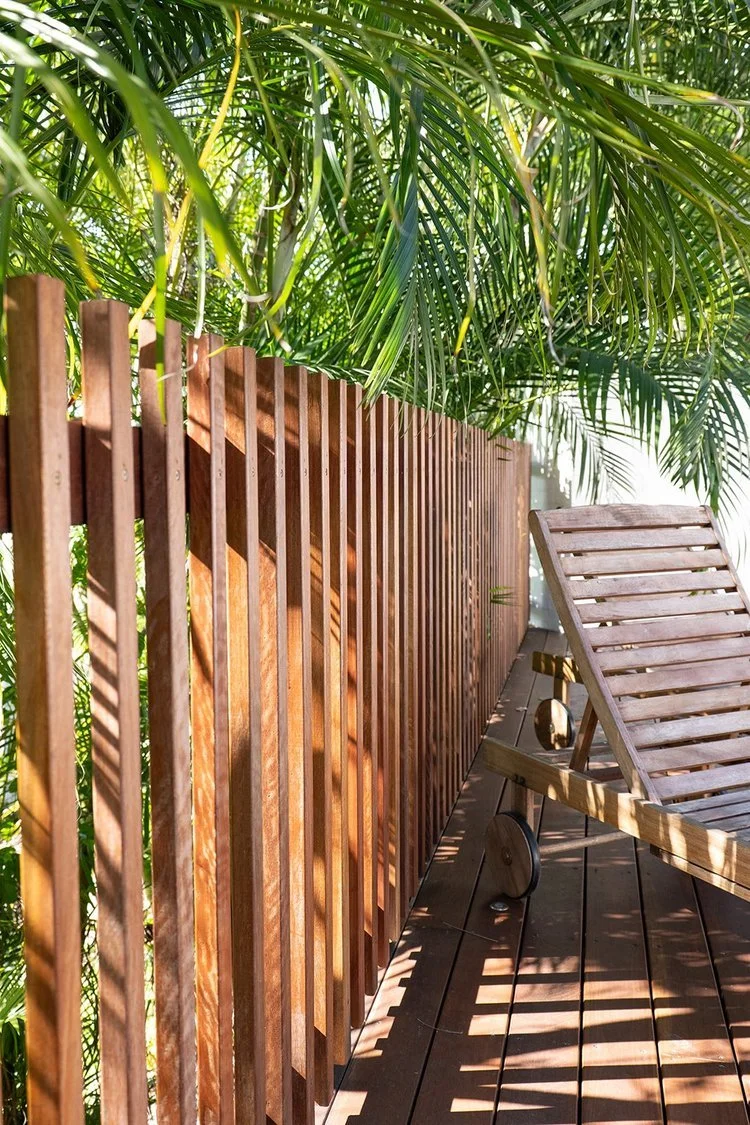 Wooden deck with a slatted wooden lounge chair, and a wooden fence with vertical slats, overgrown by tropical palm leaves.