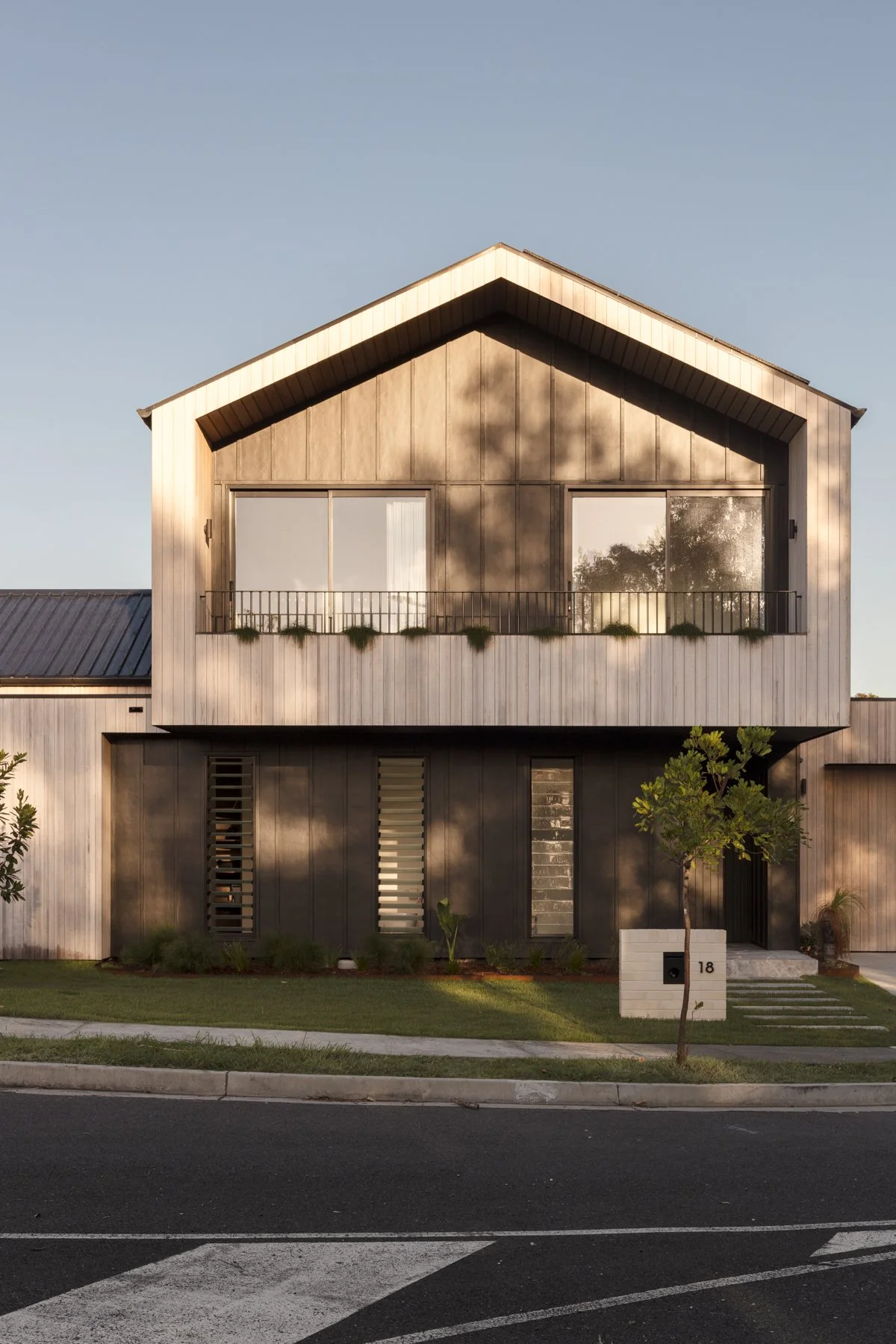 Modern two-story house with a wooden exterior, large windows, and a small balcony. Front yard with a small tree and neatly trimmed grass.