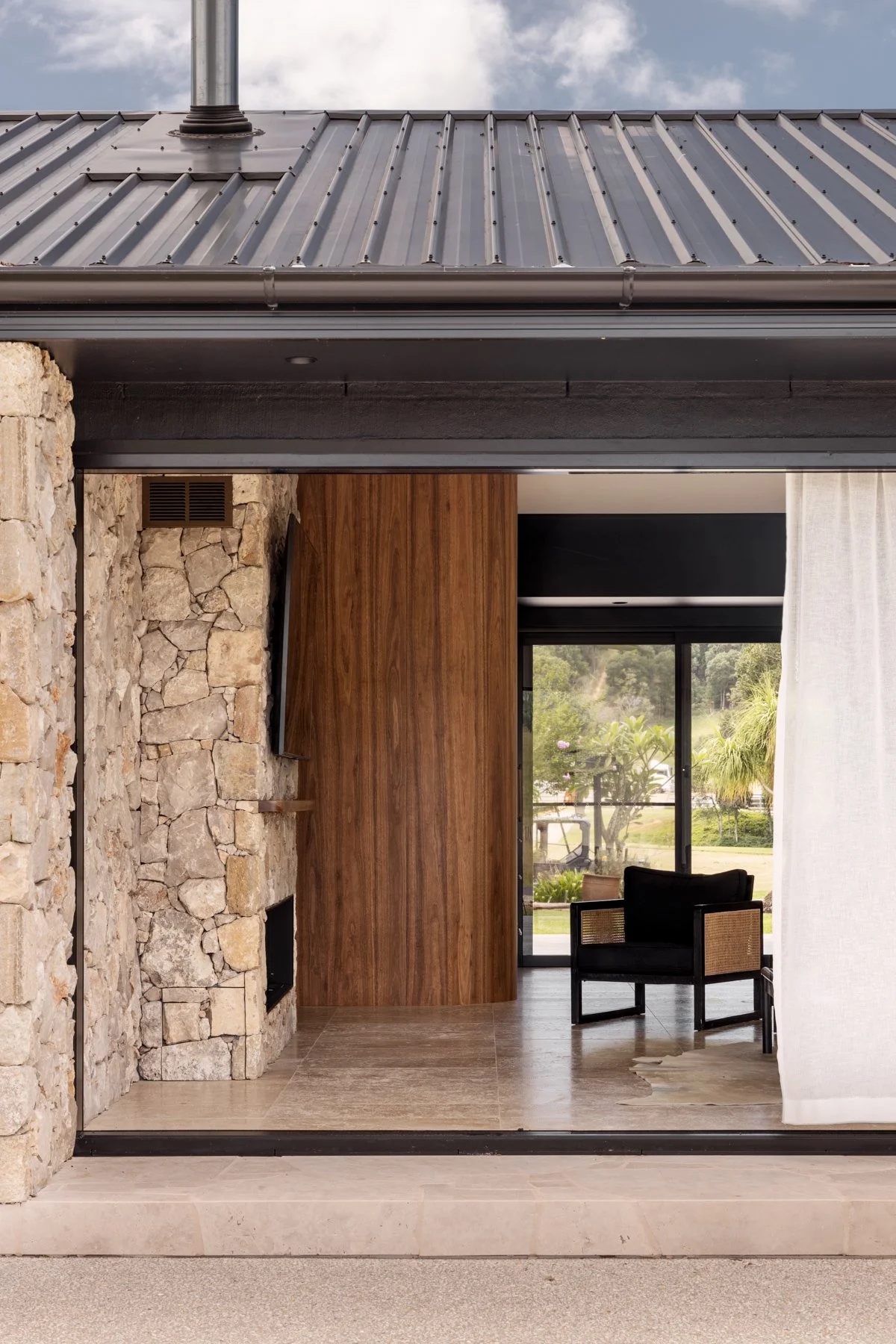 Modern house interior with stone wall, wooden panel, and glass sliding door showing outdoor greenery and seating area.