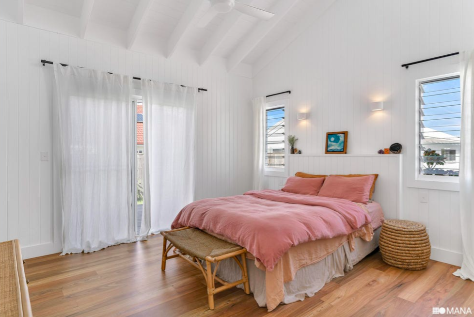 A bright bedroom with vaulted white ceiling, white paneled walls, hardwood floor, pink bedding, and sheer white curtains on sliding glass doors and windows. There is a wicker bench at the foot of the bed, a small round wicker stool on the right, and wall-mounted lights.