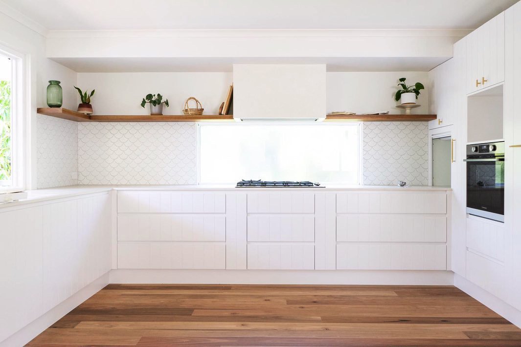 Minimalist white kitchen with wooden shelves, potted plants, and a window, featuring white cabinets and a built-in oven.