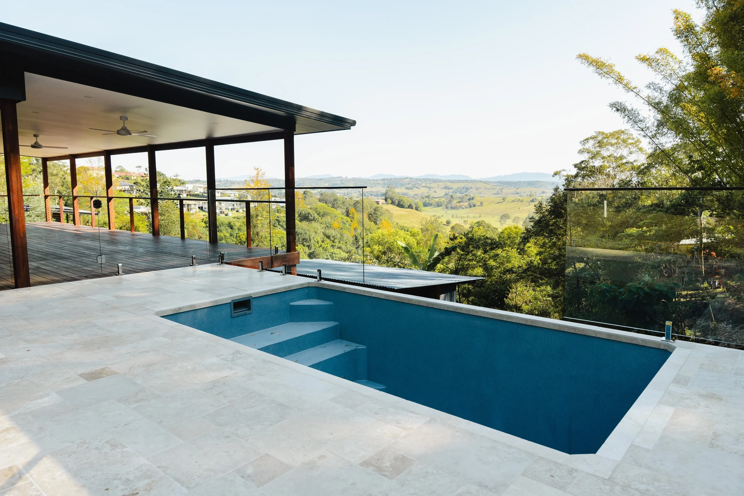 Modern outdoor balcony with glass railings overlooking a green hilly landscape with trees and distant hills on a clear day.