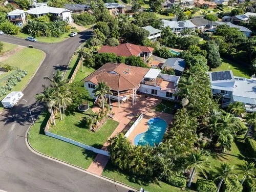 Aerial view of a large suburban house with a swimming pool, surrounded by lush greenery and palm trees, with neighboring houses and streets nearby.