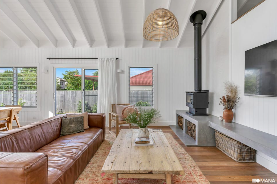 Living room with white walls, wood ceiling beams, large windows, a brown leather sofa, a beige armchair, a wooden coffee table with a plant, a wood stove, a large flat-screen TV, and a woven basket.