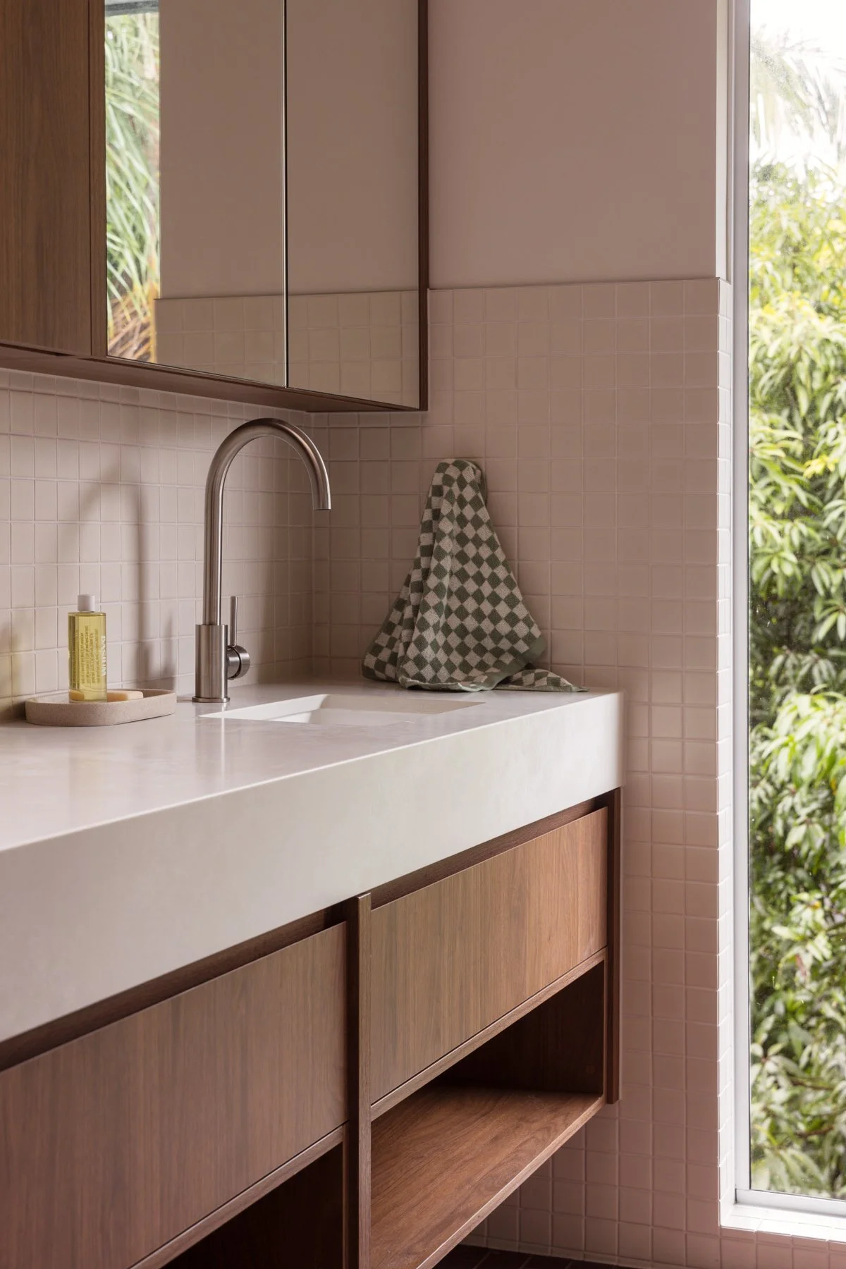 A kitchen with a white countertop, a stainless steel faucet, a beige tiled backsplash, a green and white checkered dish towel hanging on the wall, a soap dispenser on a tray, and a large window with a view of greenery outside.