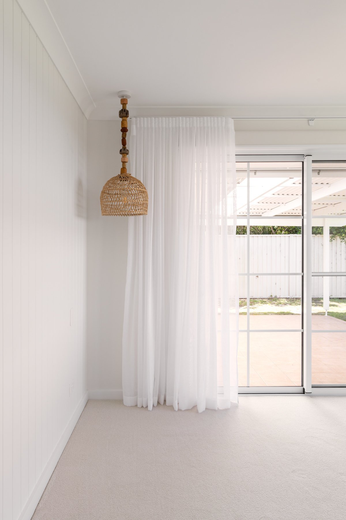 A minimalist interior room with white walls, a beige carpet, a woven rattan pendant light, white curtains, and a sliding glass door leading to a patio with a fence and some greenery outside.