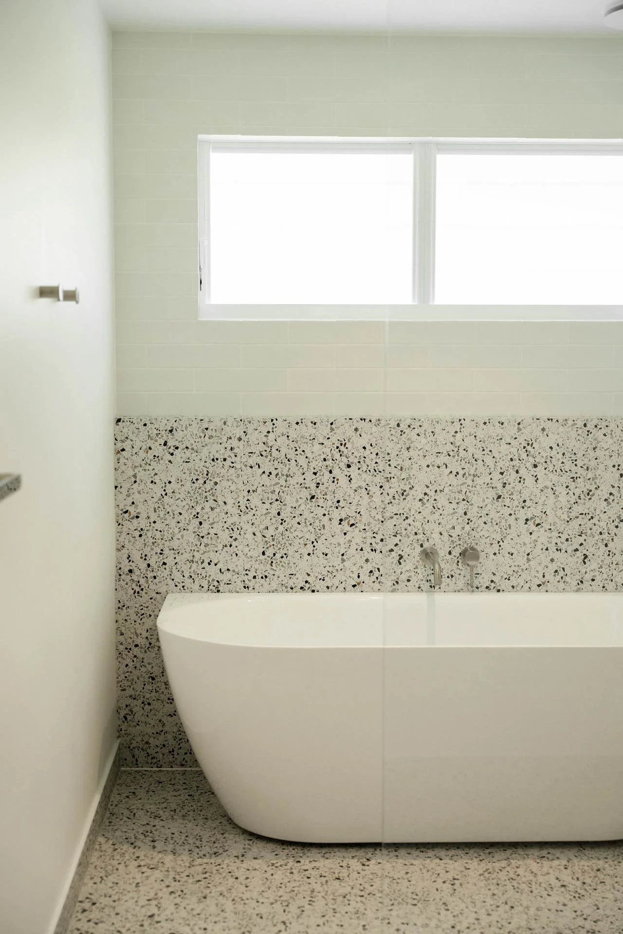 Modern bathroom with a white freestanding bathtub, terrazzo flooring, a speckled accent wall, and a large window bringing in natural light.