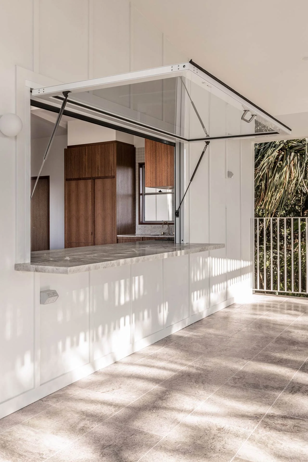 Modern kitchen with a marble countertop window opening, white walls, and a sliding window leading to an outdoor balcony with trees.