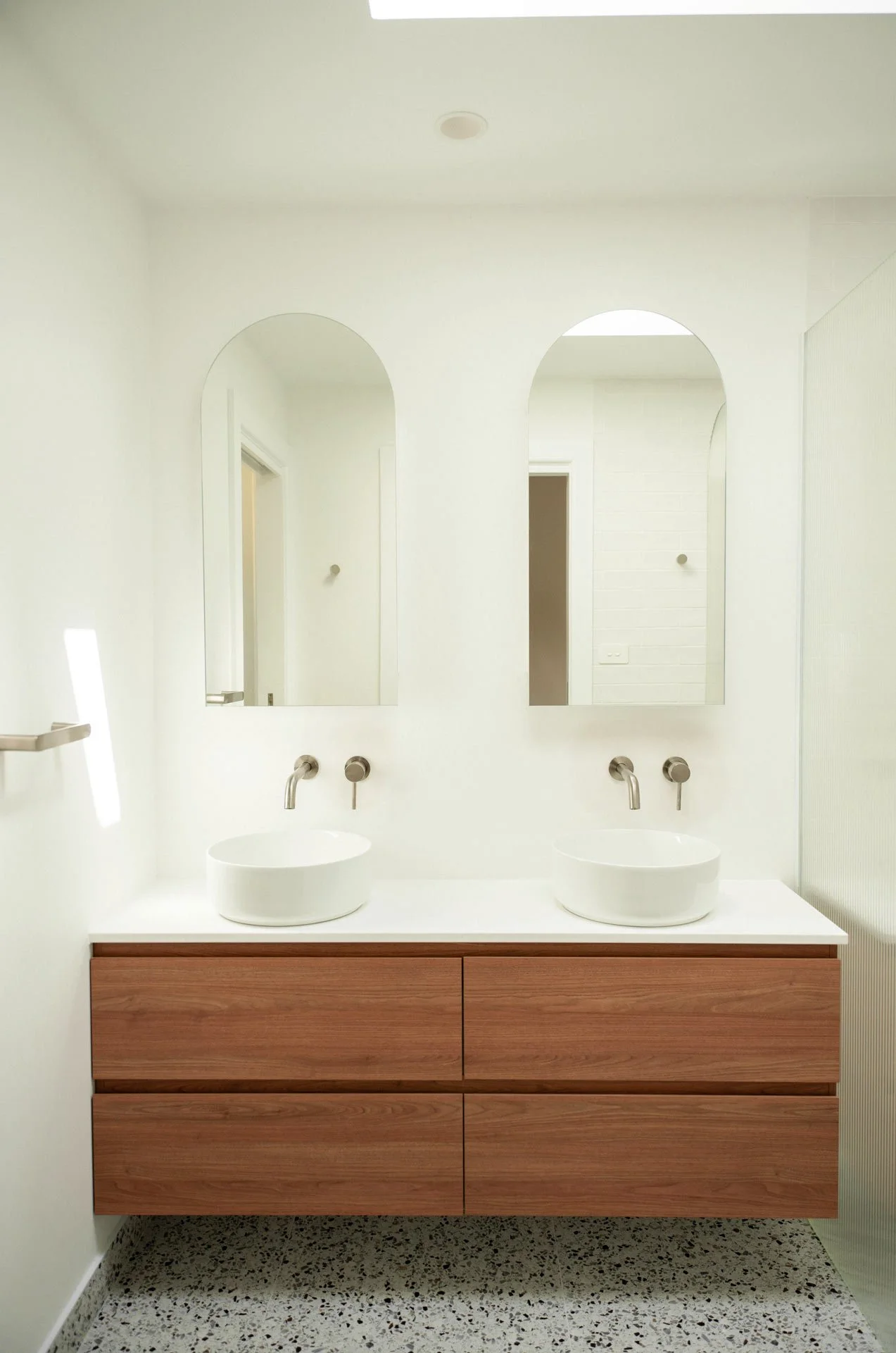 Modern bathroom with double white vessel sinks, two mirrors, and wooden vanity with drawers, terrazzo floor, and neutral-colored walls.