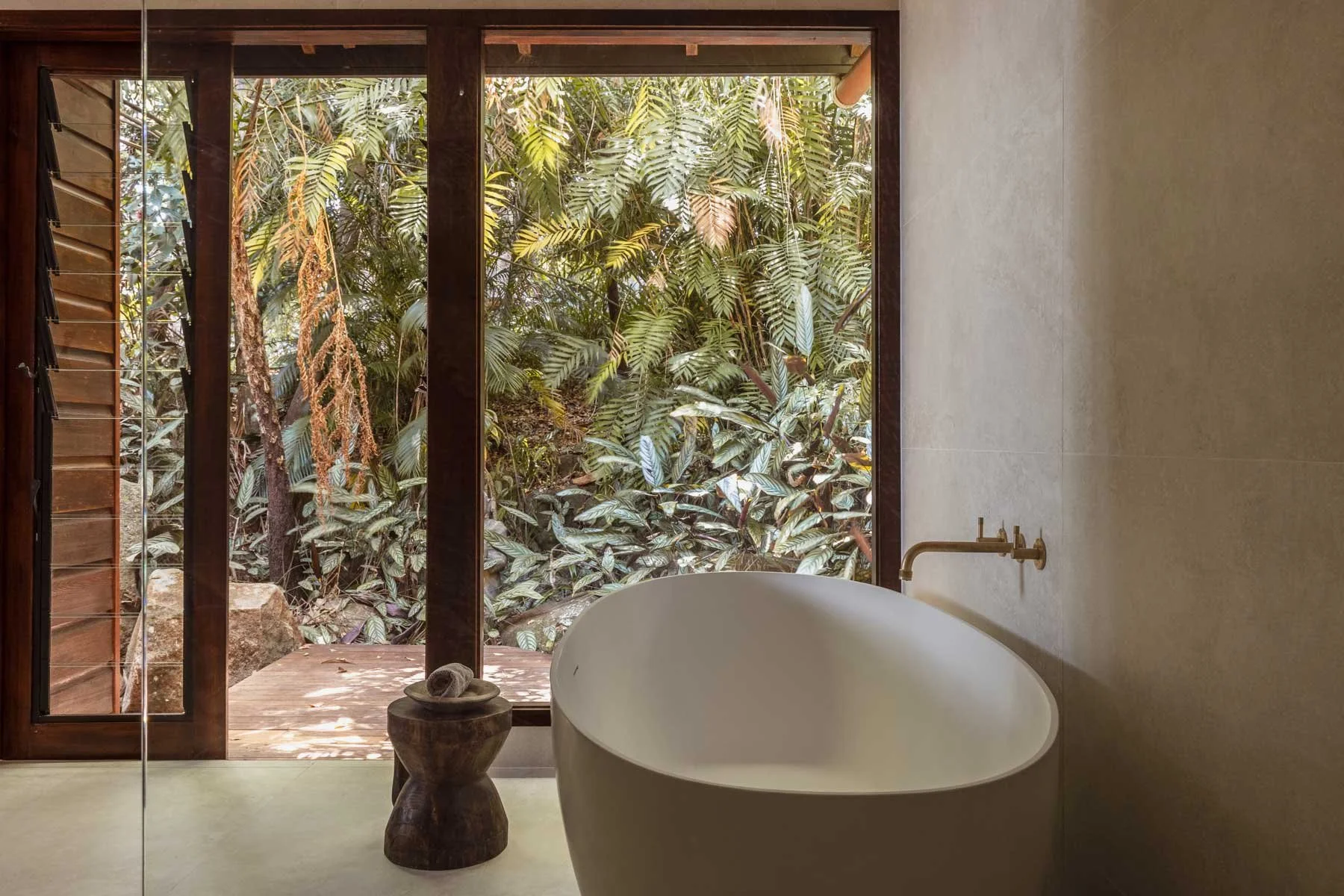 Interior of a bathroom with a freestanding bathtub facing a large window overlooking lush green tropical plants, with a small wooden side table holding rolled towels.