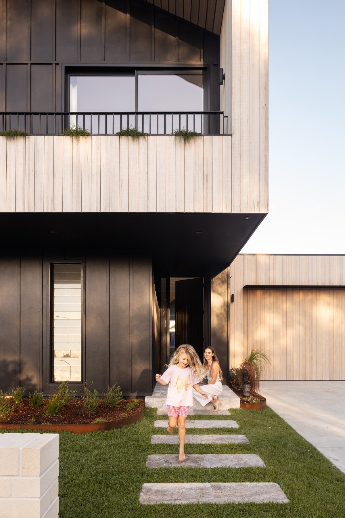 A modern house with a two-story design featuring wooden and black exterior materials, a balcony with black railing, and a front yard with a pathway. Two children are playing outside, one running and the other sitting on steps, with a woman nearby smiling.