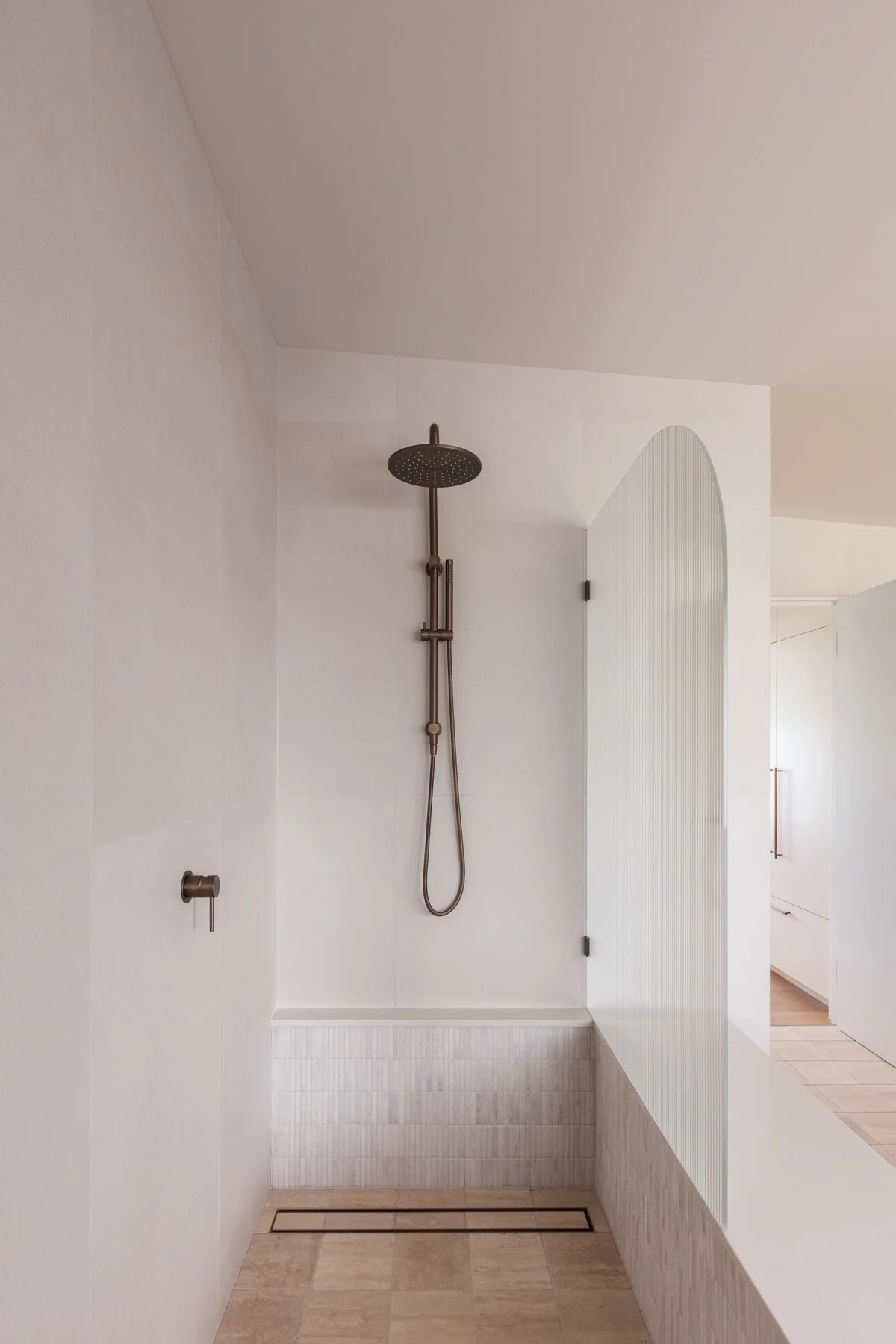 Minimalist walk-in shower with a dark bronze rain showerhead, handheld shower, and a built-in drain on light beige tiled floor with white walls.