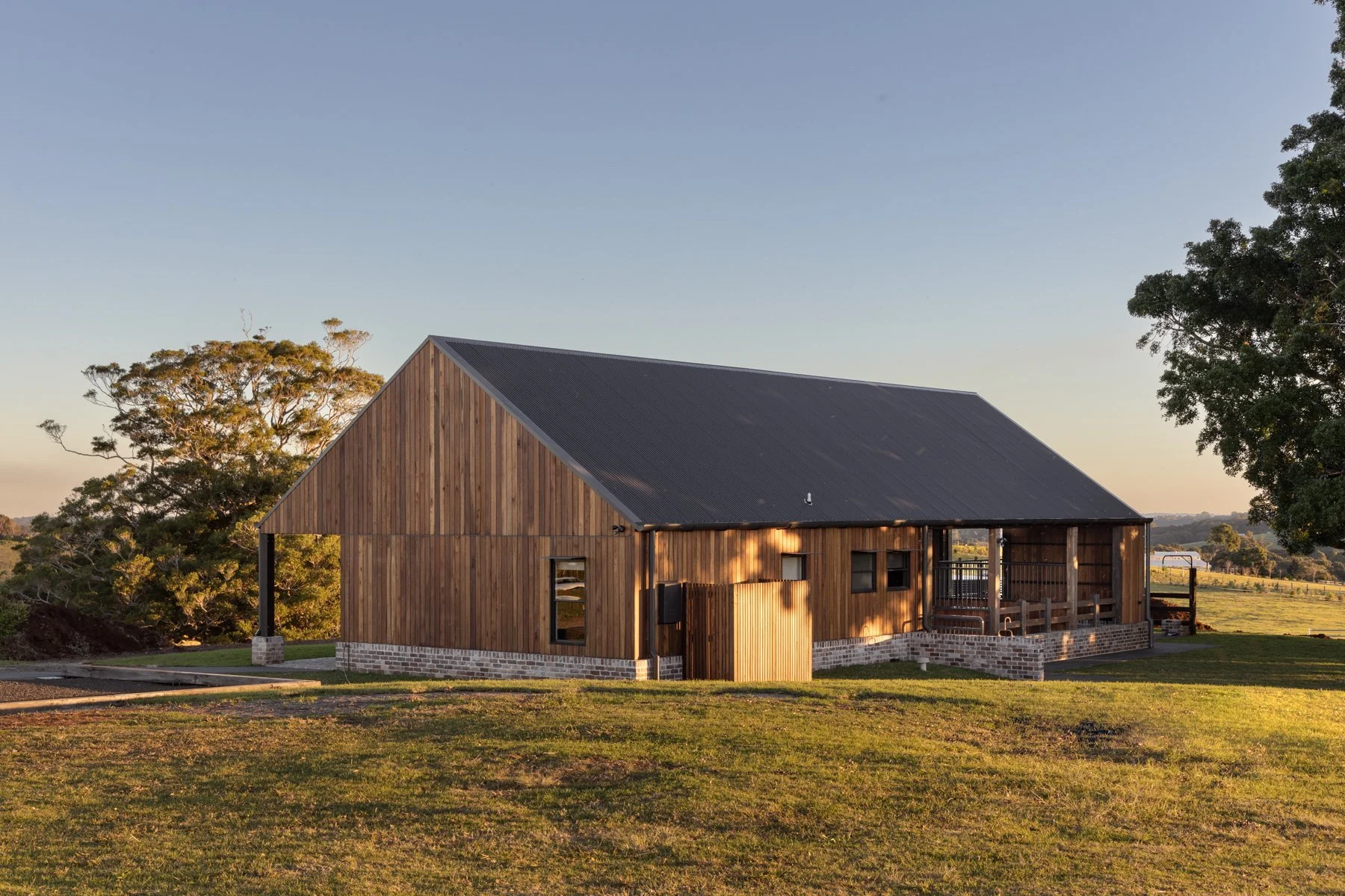 A wooden house with a metal roof, situated on a grassy area with large trees in the background, during sunset.