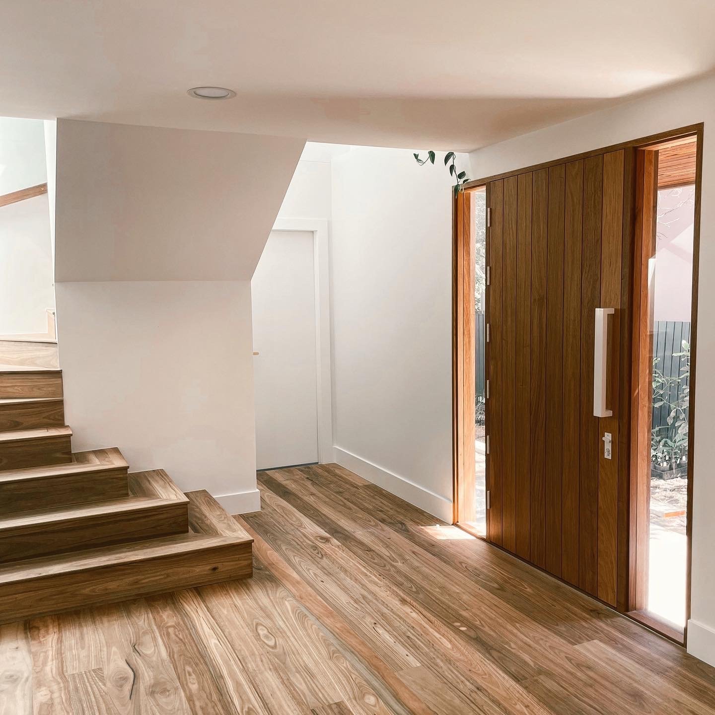 Interior view of a home’s entryway with wooden flooring, an open wooden front door, and a staircase with wooden steps to the left. There's a staircase and a small window near the door, and a plant hanging above the door.