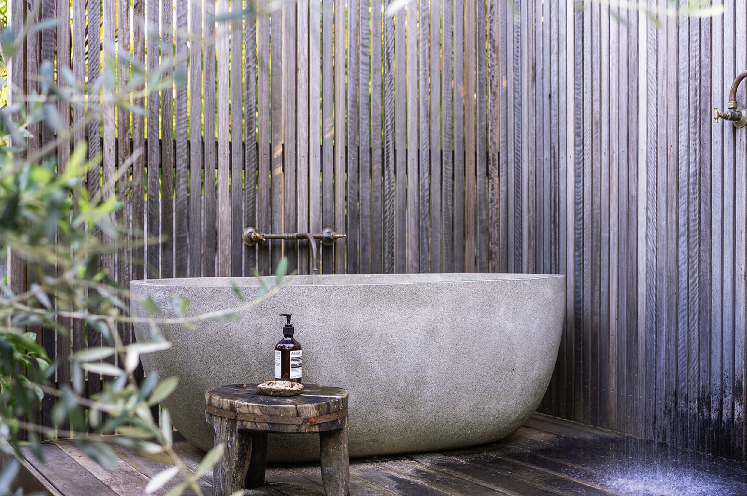 Outdoor bathtub with faucet and bottle of soap, set against a wooden privacy fence, with greenery in foreground.