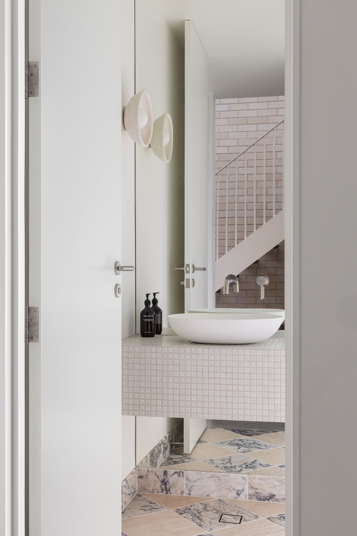Modern bathroom with white vessel sink on a tiled counter, black soap dispensers, a large mirror, and a staircase with brick wall in the background.