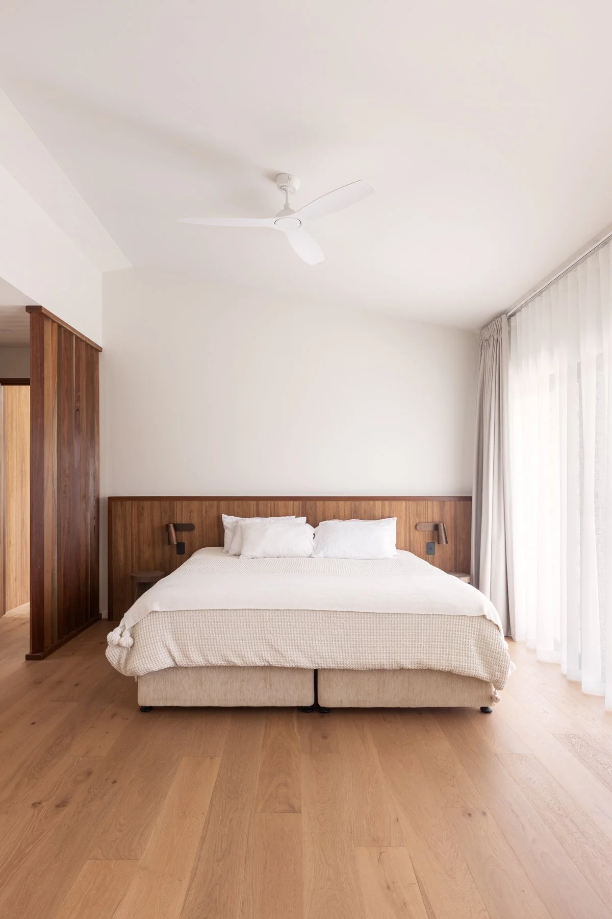 A minimalist bedroom with a bed featuring a wooden headboard and white bedding. There are two pillows, bedside lamps, and a large window with sheer curtains letting in natural light. A ceiling fan hangs from the white ceiling.