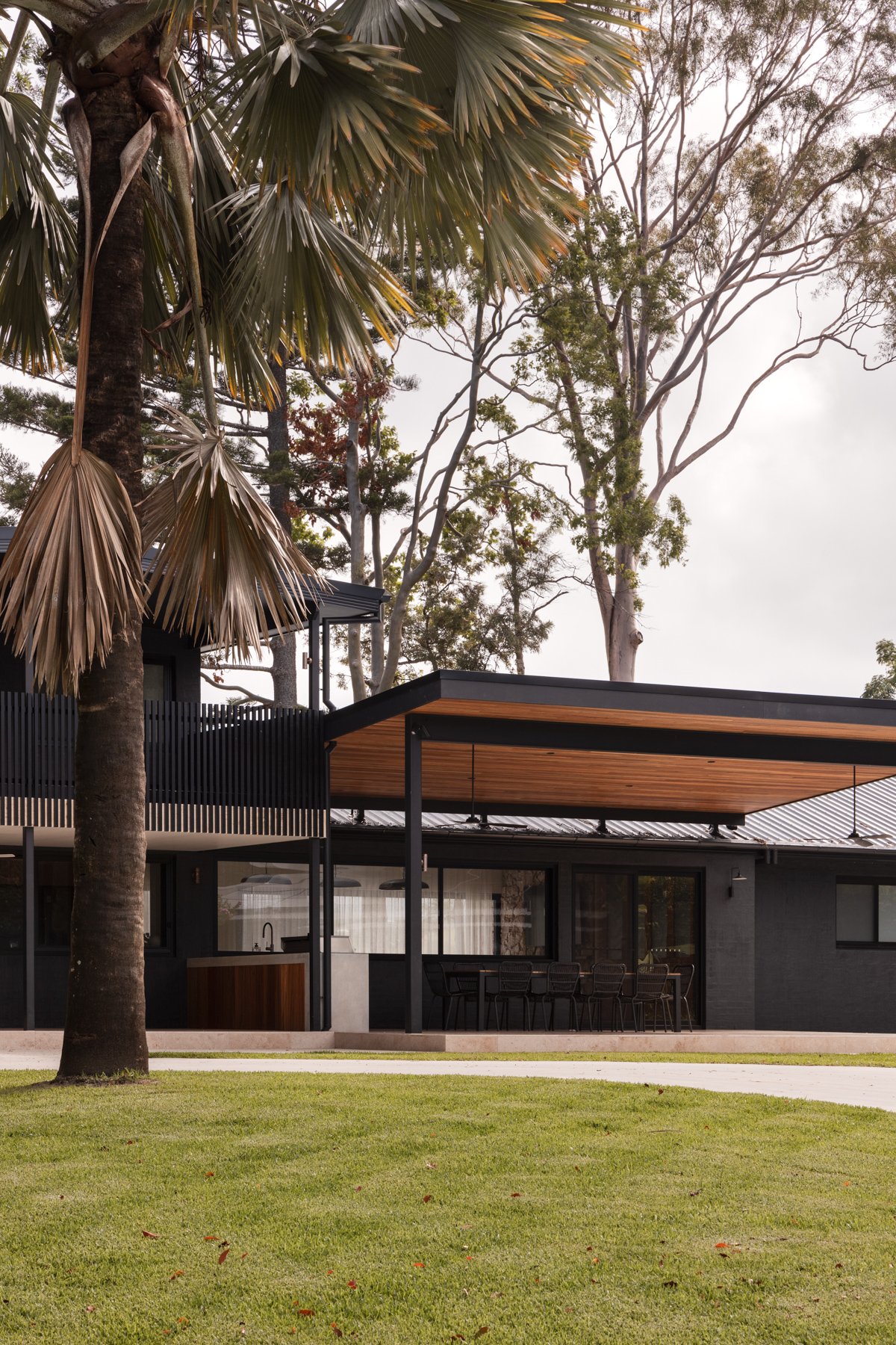 Modern house with outdoor patio, surrounded by trees and lush green lawn, under a cloudy sky.