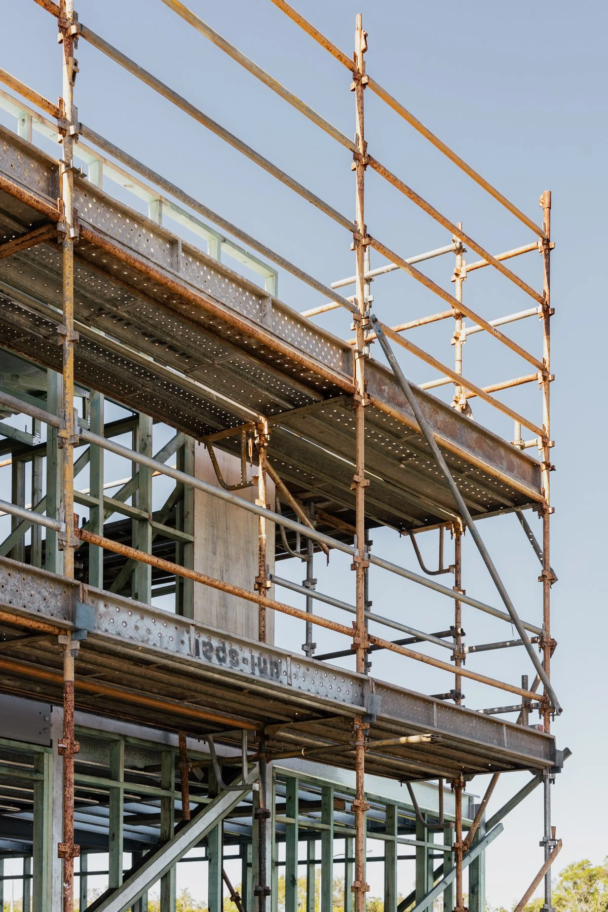 Construction scaffolding surrounding a building under construction, with metal and rusted poles and partial framing against a clear blue sky.