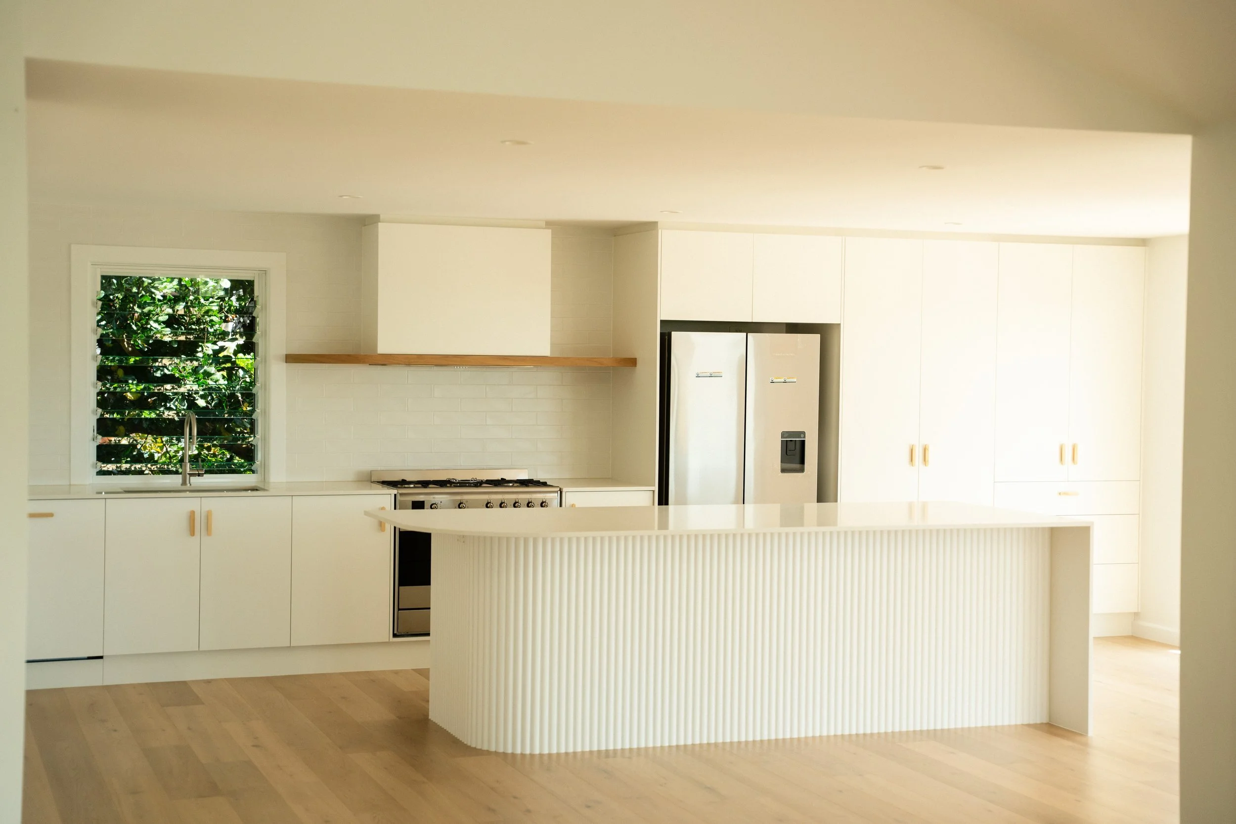 Modern kitchen with white cabinets, a white island, stainless steel refrigerator, a window with foliage outside, and light wood flooring.