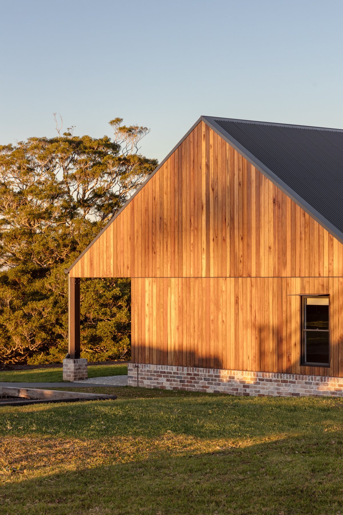 A modern house with wooden vertical siding and a black metal roof, situated on a grassy lawn with trees in the background, during sunset.