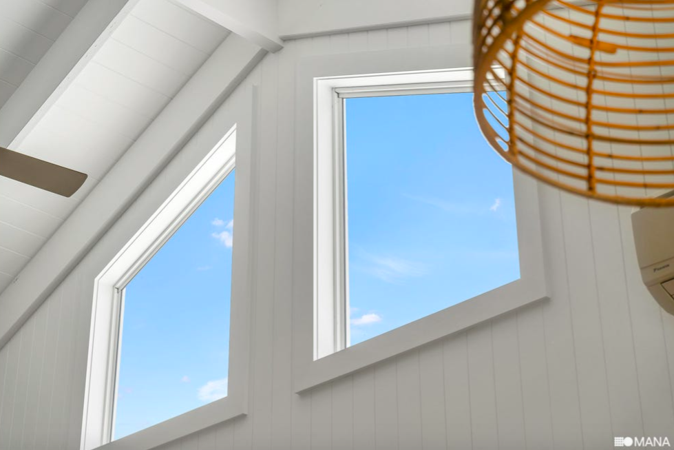 View of a bright blue sky with a few clouds seen through two white-framed pentagon-shaped windows in a room with white walls and ceiling, with part of a woven rattan light fixture visible.