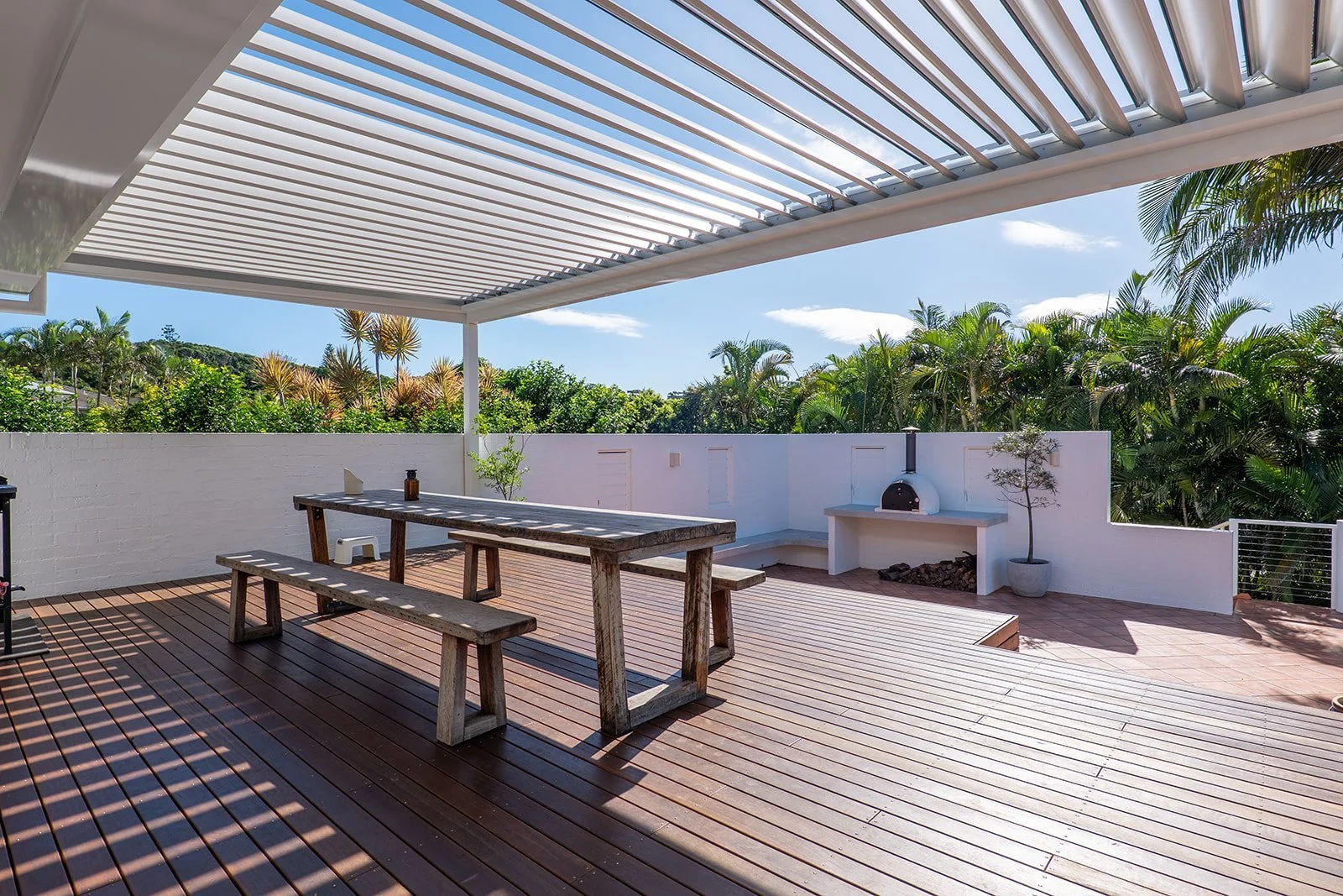 A spacious outdoor patio with wooden flooring, a rustic wooden picnic table with benches, a white brick wall with small plants, a built-in outdoor fireplace, and lush greenery and palm trees beyond the wall. The patio is shaded by a white pergola.