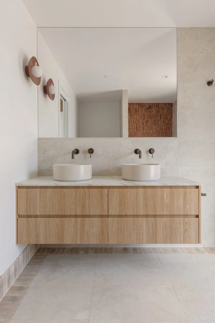 Modern bathroom with double vanity featuring wooden cabinets, two vessel sinks, and wall-mounted faucets. Large mirror and wall sconces above the sinks, with neutral-colored tile flooring and wall tiles. A glimpse of a shower area with exposed brick wall in the background.
