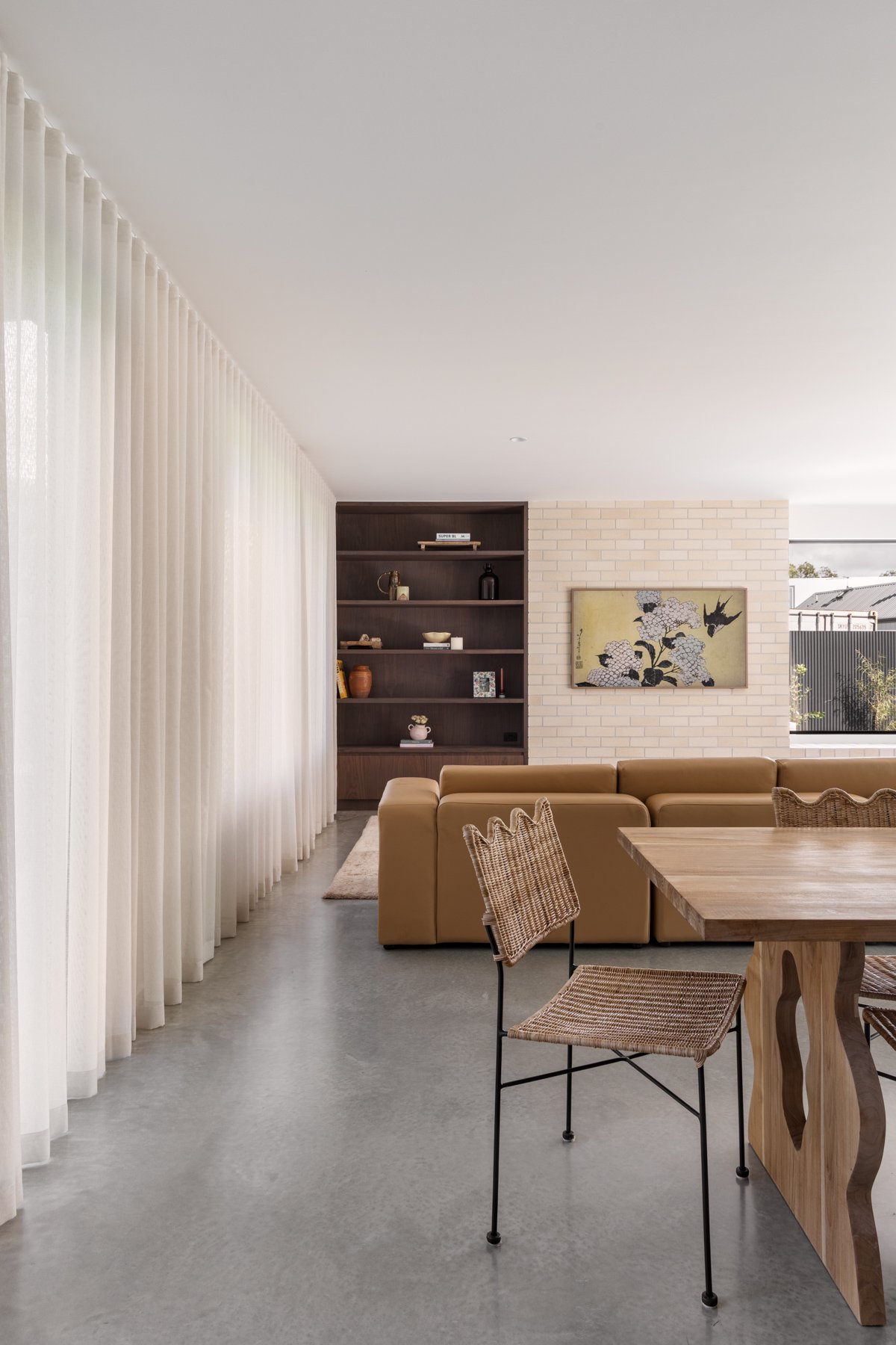 Living room with large cream sectional sofa, wooden dining table with wicker chairs, white curtains, built-in dark wood shelves with decorative items, artwork of flowers and a bird on the white brick wall, and a window showing outdoor houses.