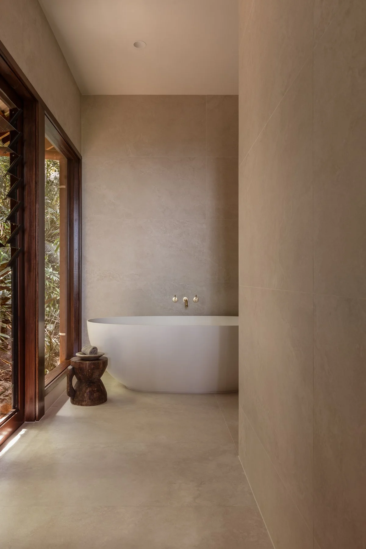 Modern bathroom with a freestanding bathtub, large window with wooden frame, beige tiled walls and floor, small wooden stool with a rolled towel placed on it.