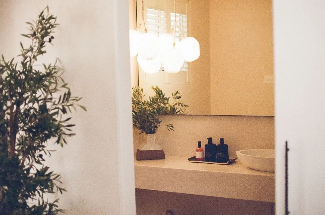 A bathroom with a large mirror, a countertop with black and pink bottles, a white bowl, a potted plant, and a window with blinds.