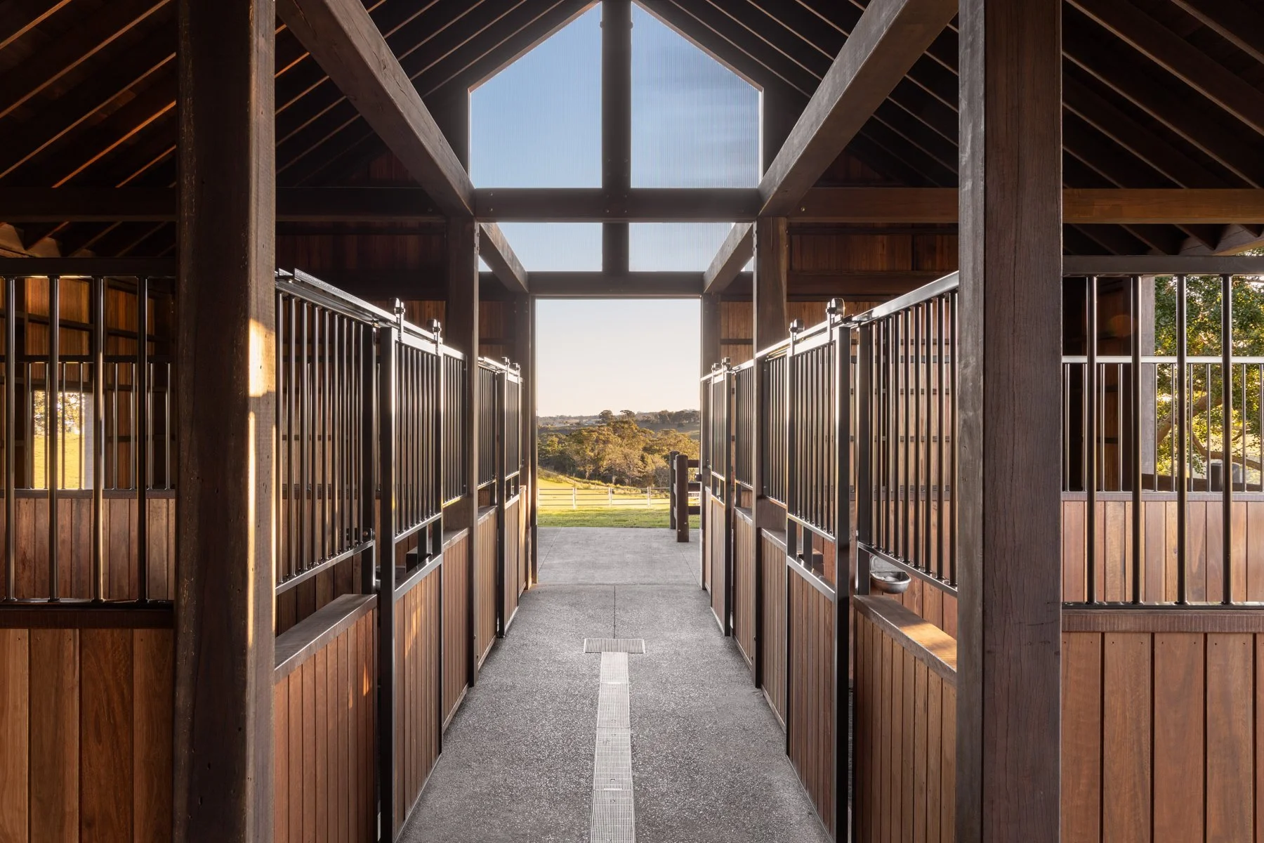 View of a horse stable corridor with wooden stalls and black metal bars, leading to an outdoor landscape with trees and a clear sky at sunset.