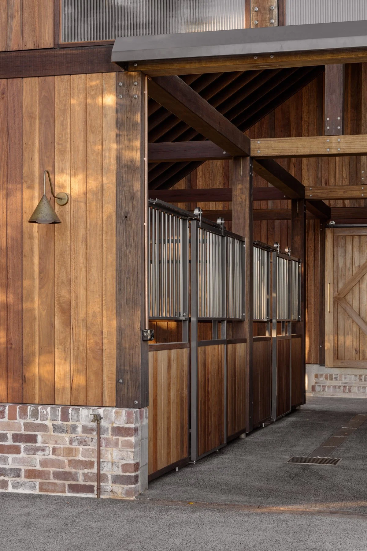 Exterior of a rustic stable with wooden walls, metal gates, and a small wall-mounted lamp.