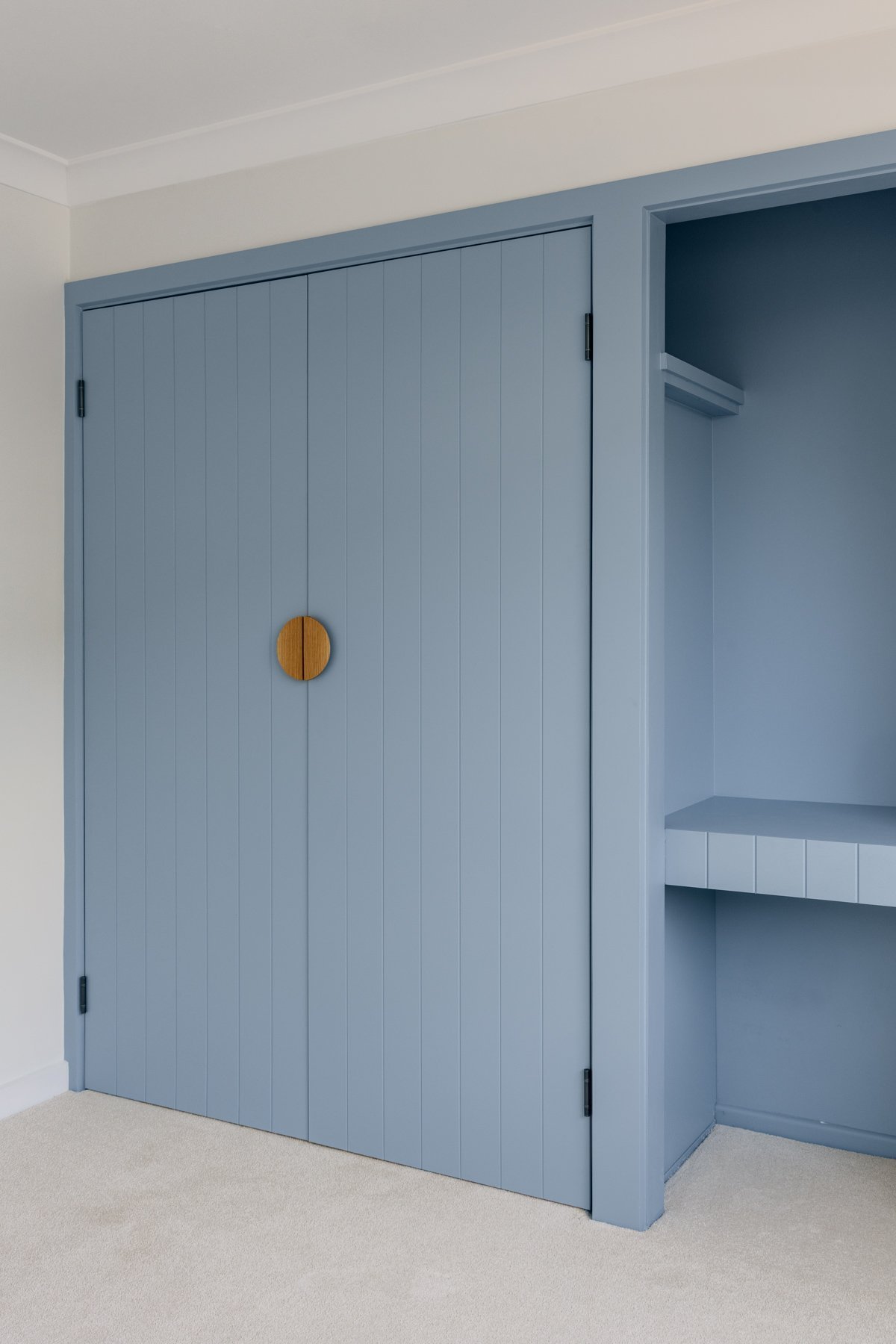 Blue built-in closet with double doors, round wooden door pull, and an open side shelf with a bench underneath, inside a room with light-colored carpet and white walls.