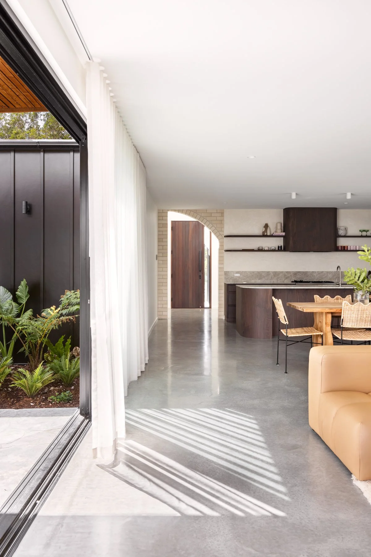 Open sliding glass door leading to a modern interior living space with a kitchen and dining area, sunlight casting striped shadows on polished concrete floor, plants outside, and a wooden front door in the background.